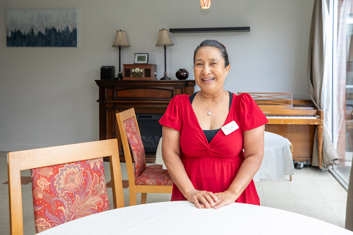 A woman in a red dress smiles warmly while standing by a table in a cozy indoor setting.