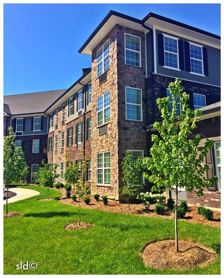 A modern apartment building with a brick facade, surrounded by neatly landscaped lawns and young trees on a sunny day.
