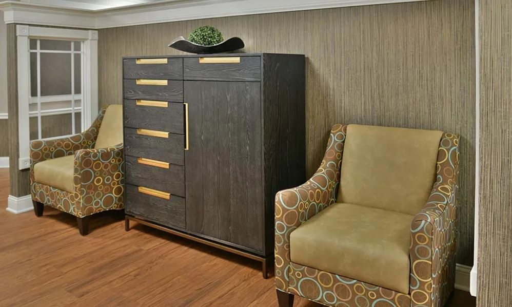 A modern interior features a wooden cabinet with decorative handles, flanked by two patterned armchairs on a wood floor.