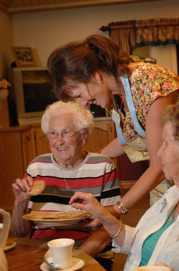 A caregiver assists elderly individuals during a meal in a communal setting.