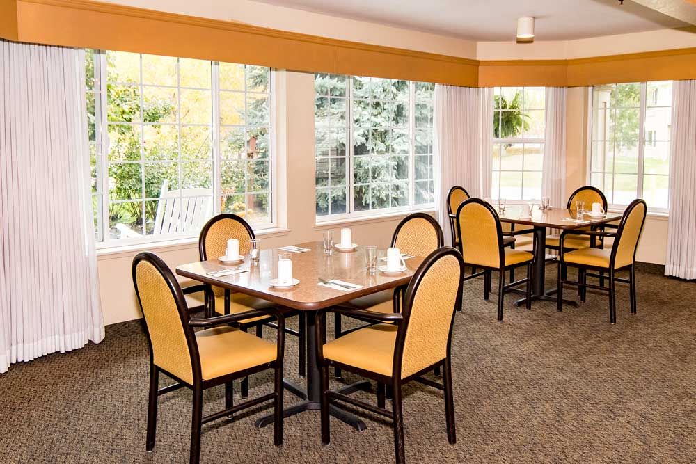 A cozy dining room with neatly arranged tables and chairs, illuminated by natural light coming through large windows.