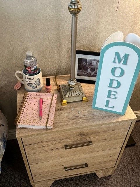 A nightstand with a lamp, notebook, pen, water bottle in a mug, nail polish, photo frame, and a 'MODEL' sign.