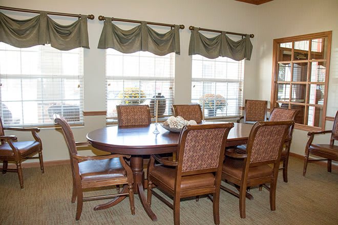 A cozy dining room with a wooden table and six chairs, framed by three large windows with green curtains.