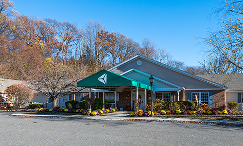 A building with a green awning is surrounded by autumn trees and plants, featuring a parking lot in the foreground.
