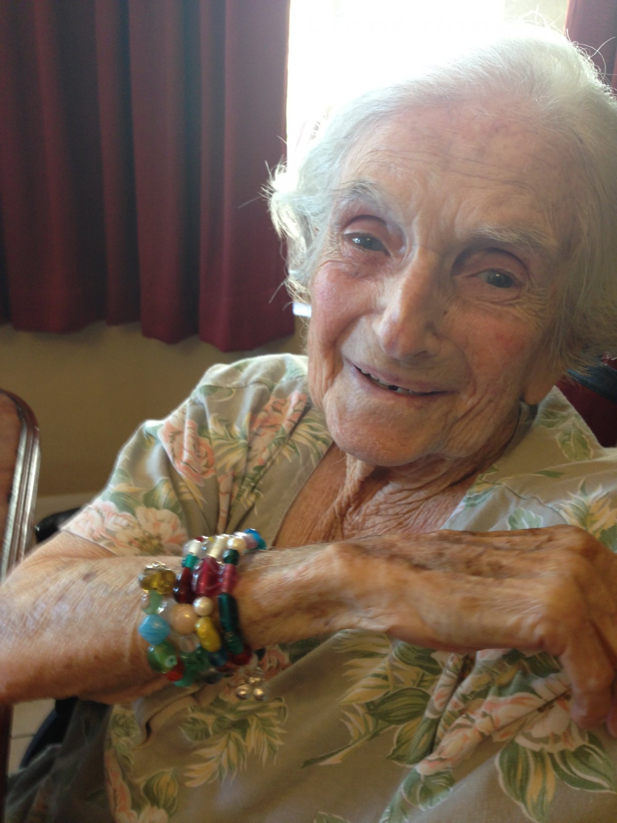 An elderly woman wearing colorful bracelets is smiling while seated indoors.