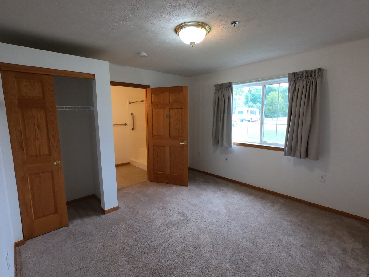 An empty bedroom with carpet flooring, a window with curtains, and an open closet door.