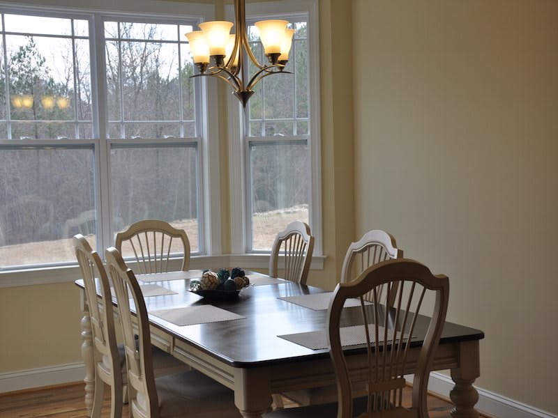 A cozy dining area with a wooden table and chairs, illuminated by a chandelier next to large windows.