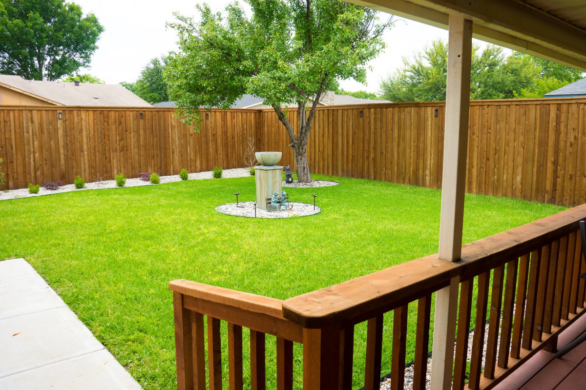 A cozy backyard with a green lawn, tree, and barbecue grill bordered by a wooden fence.
