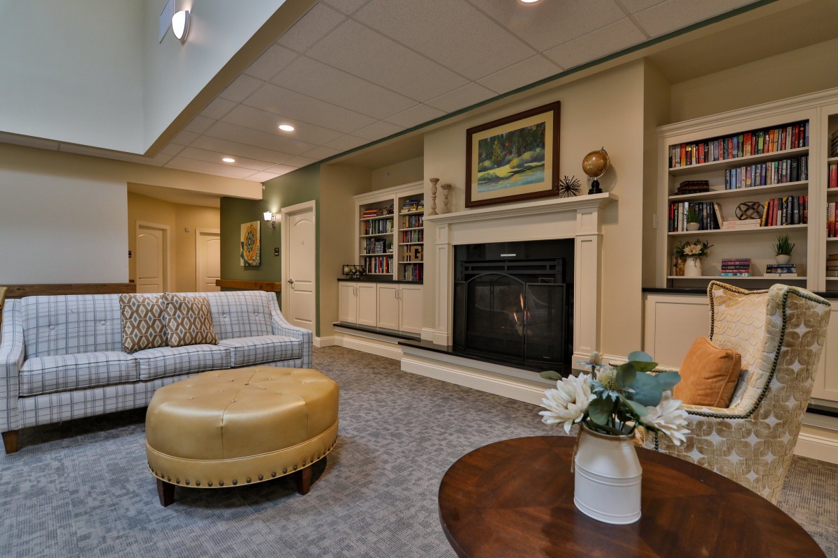 A cozy living room with a fireplace, surrounded by bookshelves and decorated with a sofa and ottoman.