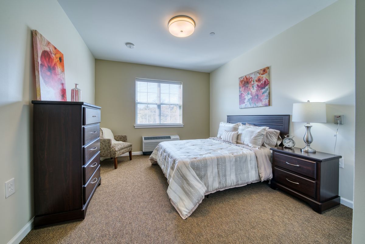 A cozy bedroom with a bed, dresser, and chair, featuring warm lighting and neutral decor.