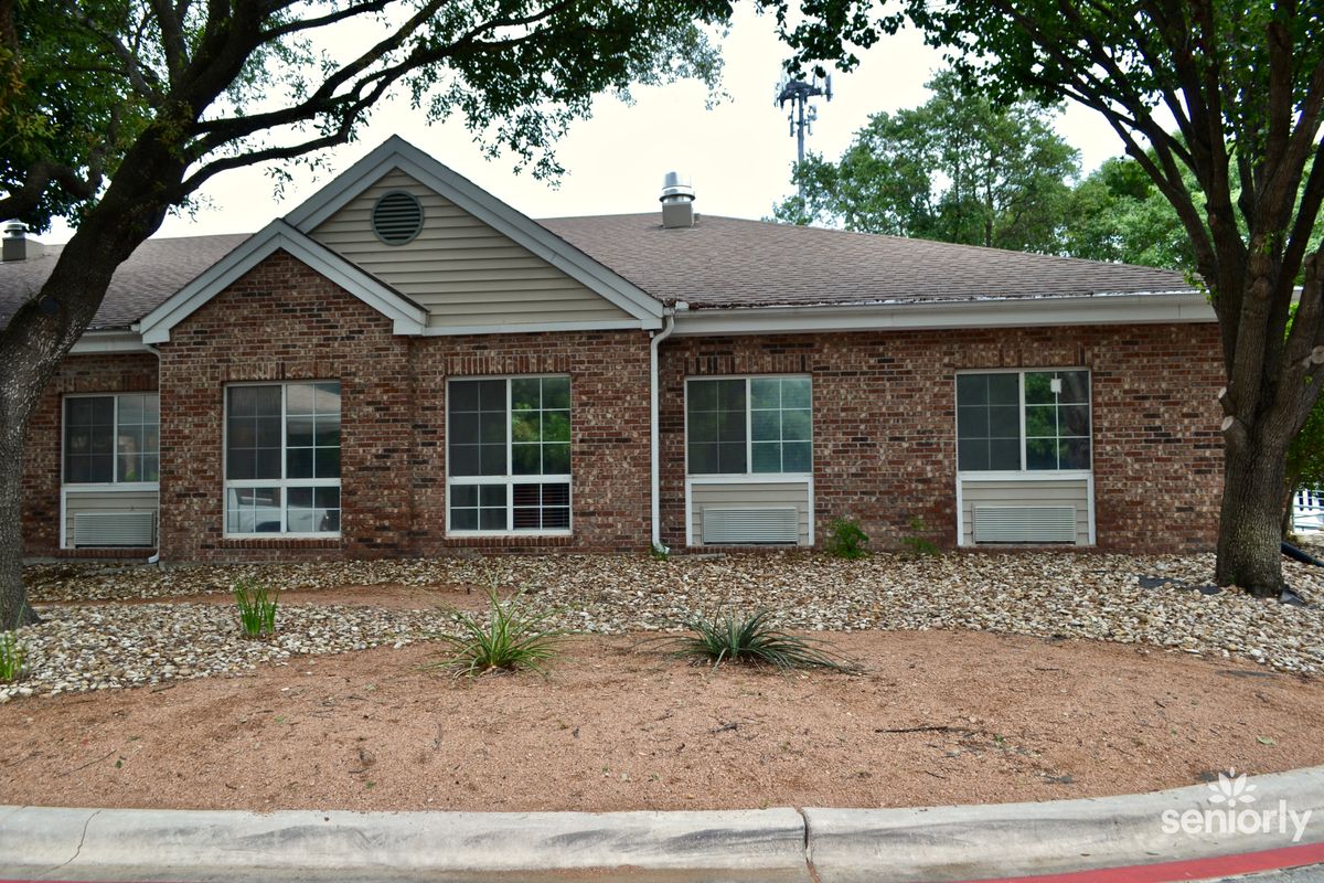 A brick residential building with large windows and minimal landscaping, featuring a row of small plants and trees in the background.