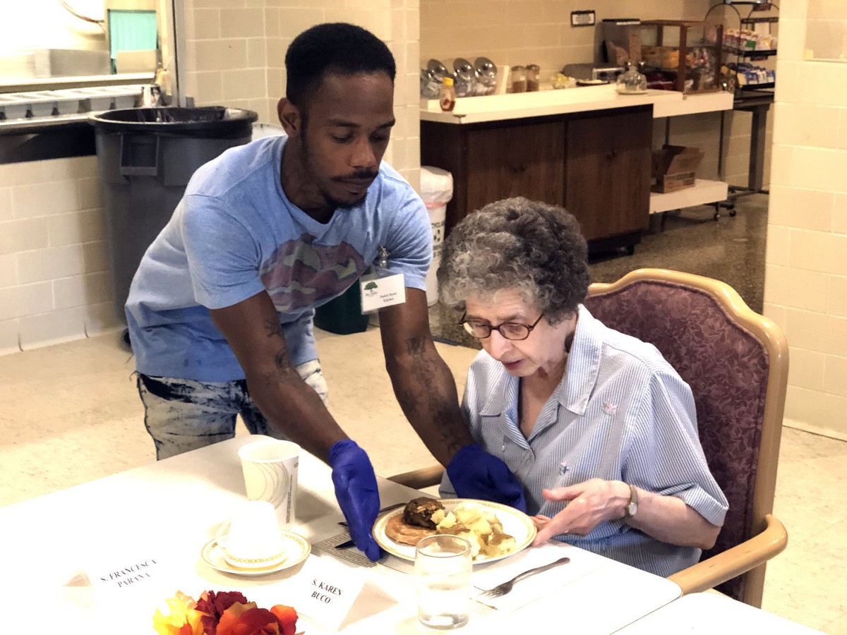 A caregiver assists an elderly woman with her meal at a dining table.