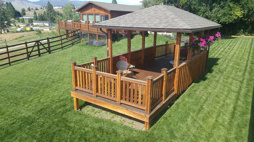 A wooden gazebo located in a backyard, featuring a table and surrounded by green grass.