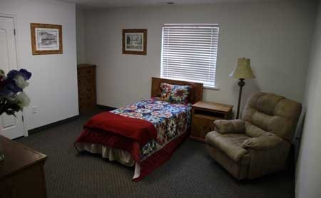 A cozy bedroom featuring a single bed with a colorful quilt, an armchair, and a lamp near the window.
