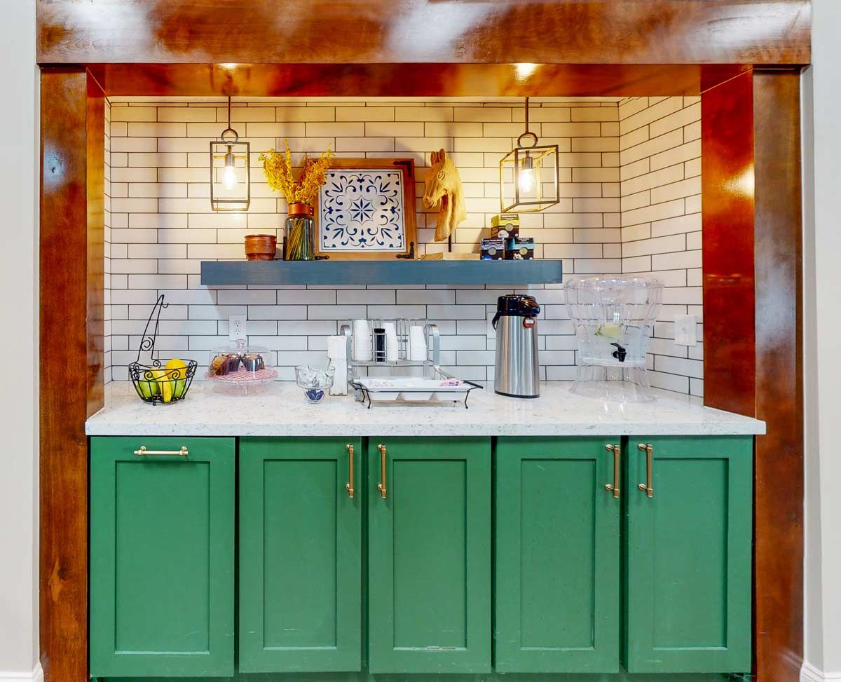 A modern kitchen with green cabinets and white subway tile backsplash, featuring a coffee setup and decorative items on a shelf under pendant lights.