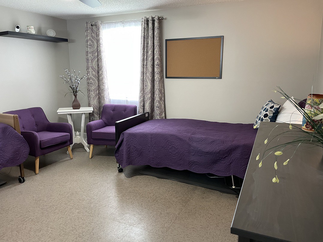 A minimalist bedroom with purple bedding, two chairs by a window, and a small table.