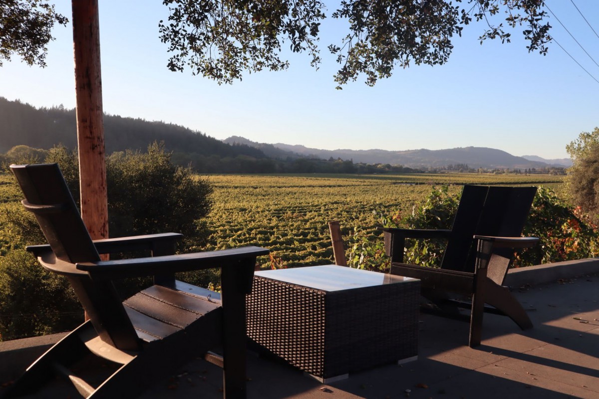 A tranquil outdoor seating area overlooking a vineyard with mountains in the background during sunset.