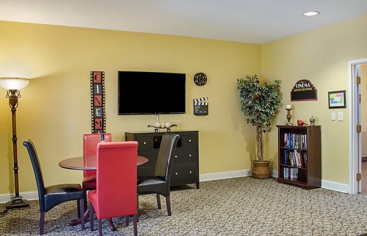 A cozy living and dining area featuring vibrant red chairs, a television mounted on the wall, a decorative clock, an indoor plant, and a bookshelf against yellow walls.