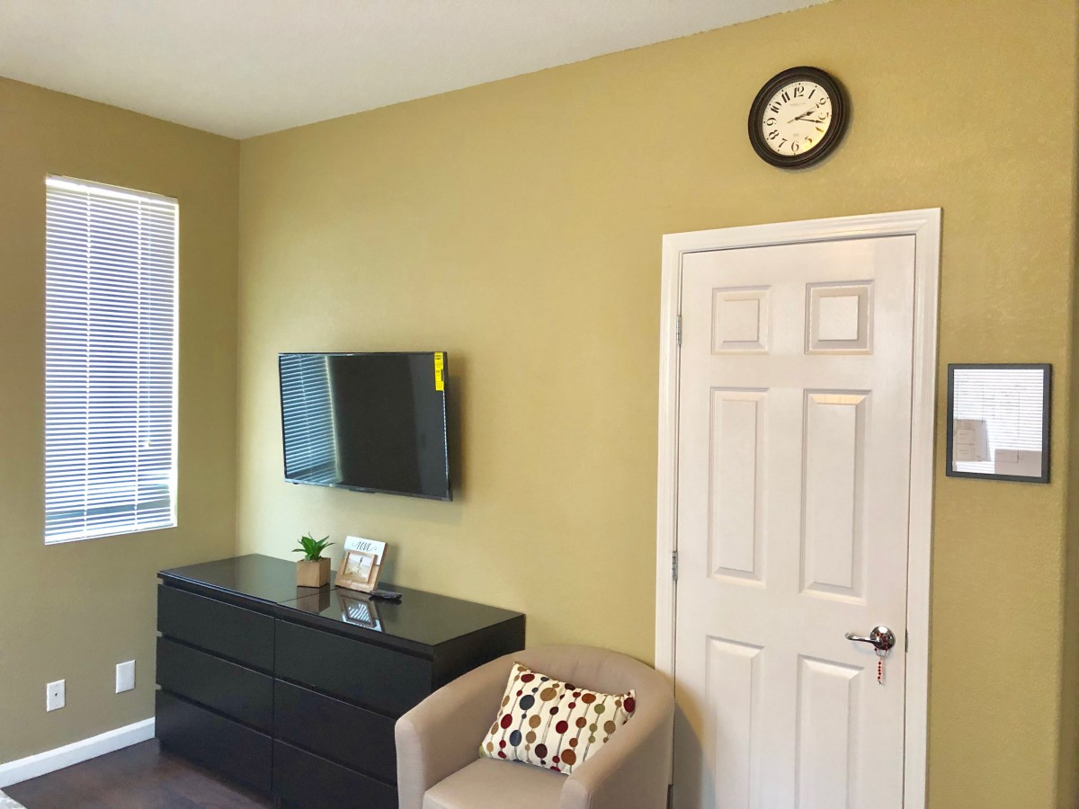 A cozy room interior featuring a wall-mounted television, a black dresser with decorative items, a comfortable chair with a patterned pillow, and a wall clock above the door.