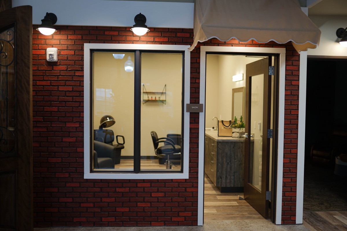 A cozy barbershop with chairs visible through the window, featuring a brick exterior and warm interior lighting.