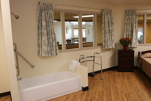 A well-lit healthcare room featuring a bed, walker, and large windows with curtains.