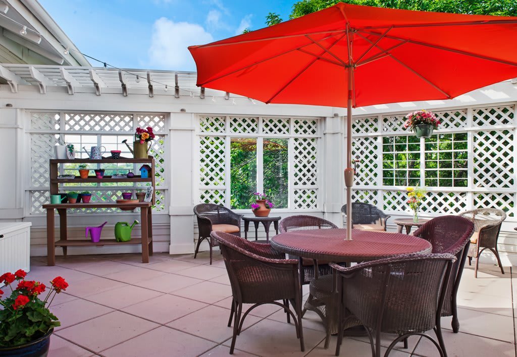 A cozy outdoor patio with a red umbrella shading a table surrounded by wicker chairs.