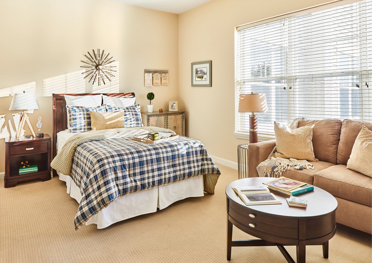 A cozy bedroom with plaid bedding, accented by a stylish chair and side table, illuminated by natural light through window blinds.