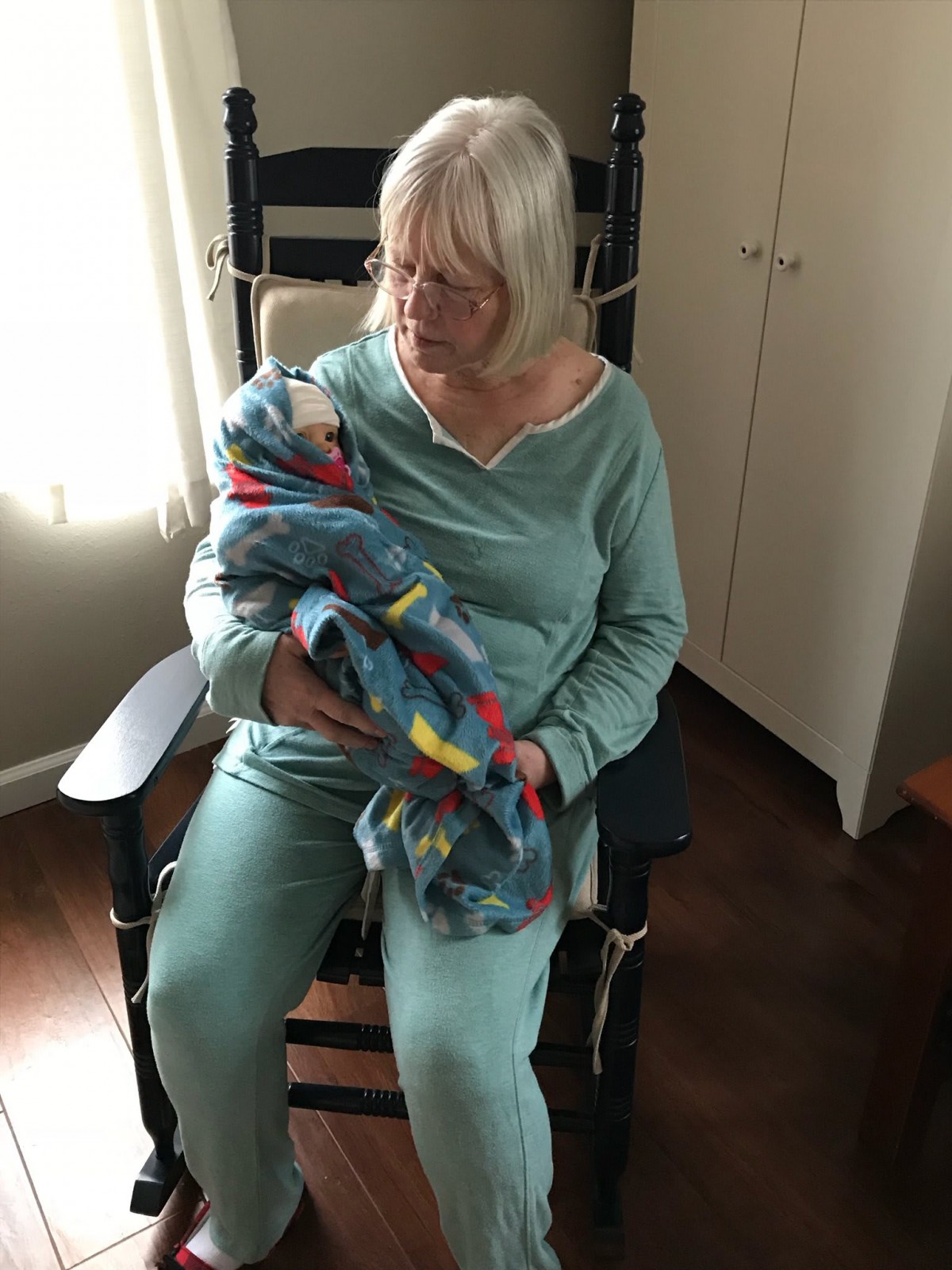 An elderly woman gently holds a baby wrapped in a colorful blanket while sitting in a rocking chair indoors.
