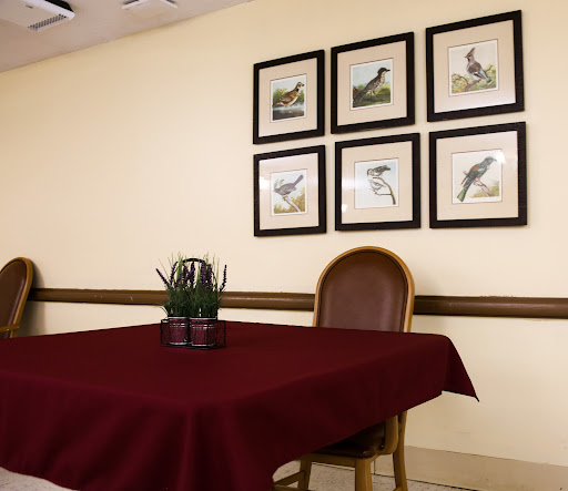 A dining room setting with a maroon tablecloth, featuring a small plant centerpiece and framed bird artwork on the wall.