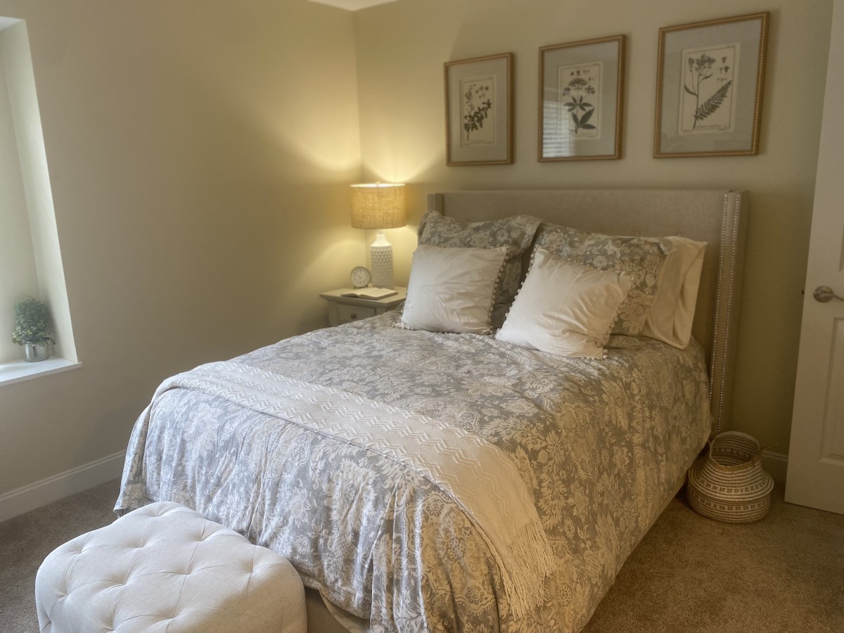 A cozy bedroom featuring a neatly made bed with patterned bedding, decorative cushions, a bedside lamp, and artwork on the wall.