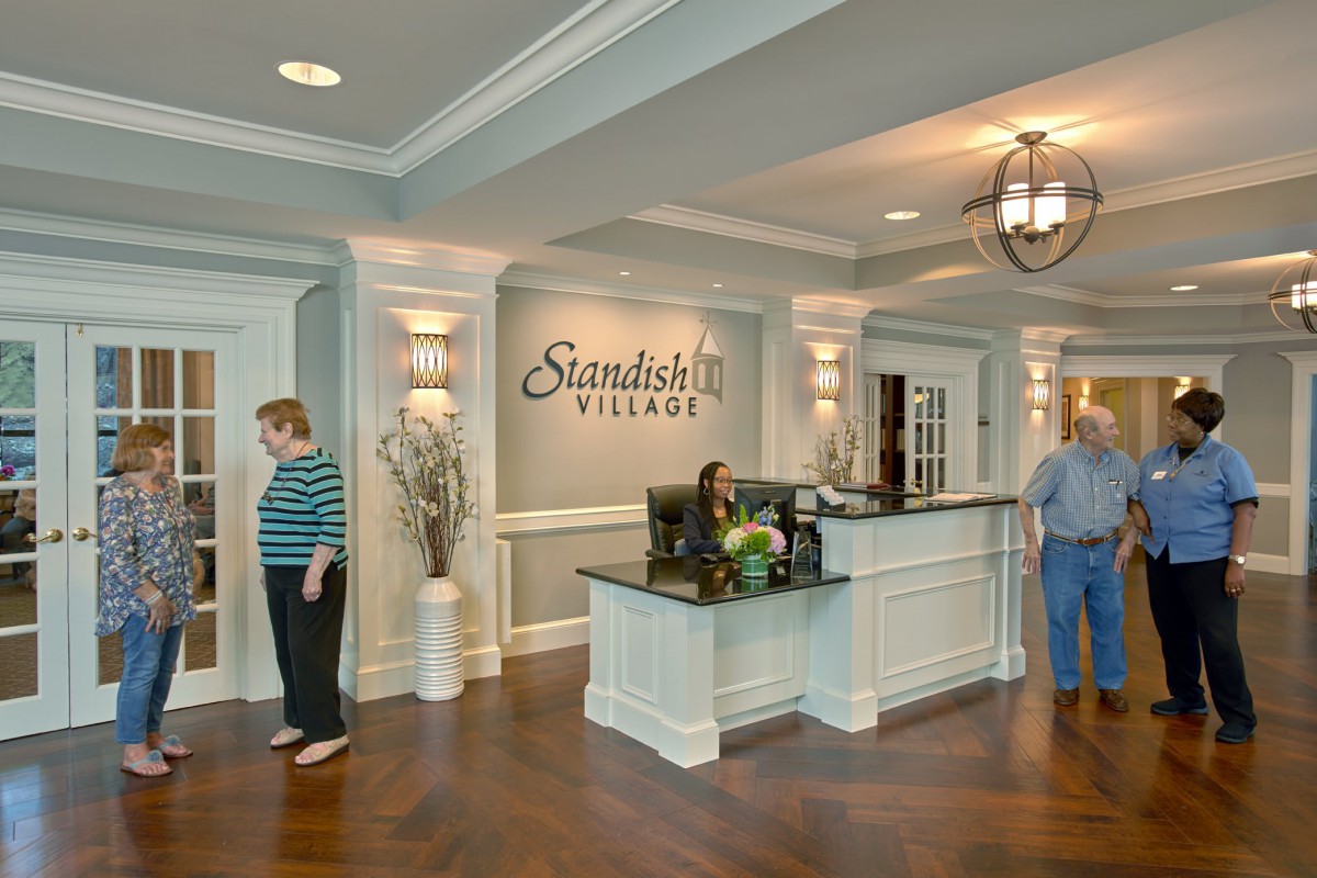 A reception area with a sign that reads 'Standish Village', featuring a front desk and several people in an elegantly designed interior.