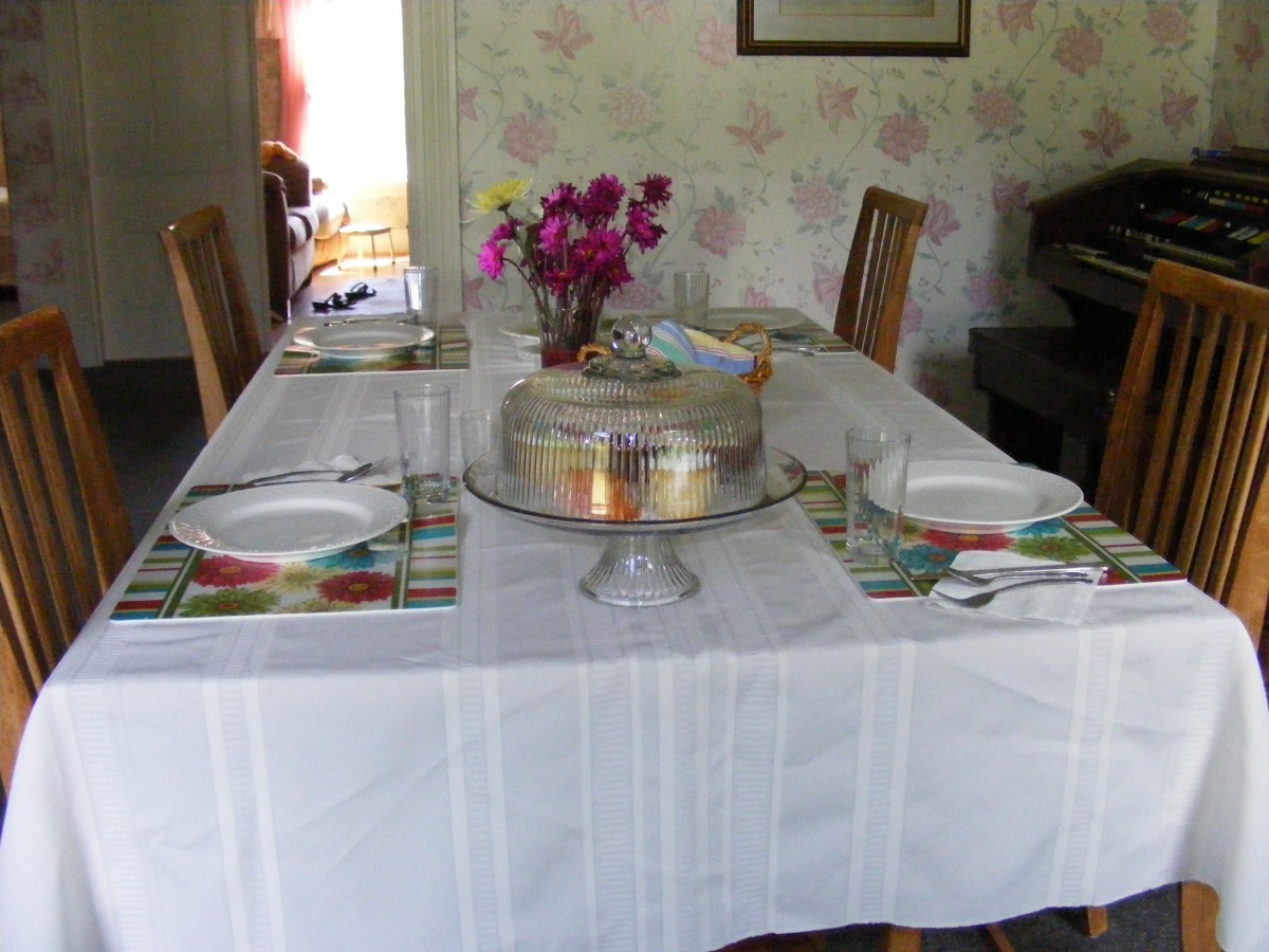 A dining table set with a white tablecloth, featuring plates, a covered cake, and a bouquet of flowers.