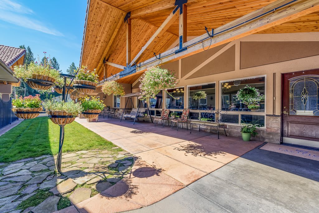 A modern building with an extended wooden roof and outdoor patio area, surrounded by greenery on a sunny day.