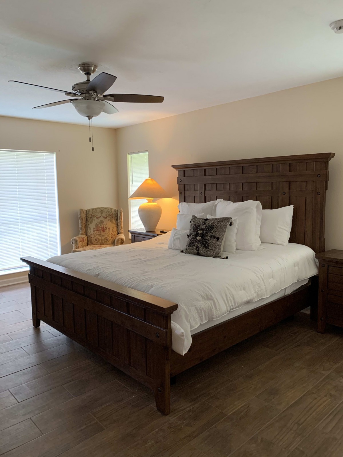 A cozy bedroom featuring a large wooden bed, ceiling fan, and soft natural lighting.