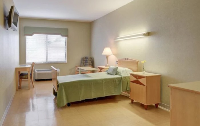 A clean and minimalist hospital room with a single bed, side tables, and a lamp.