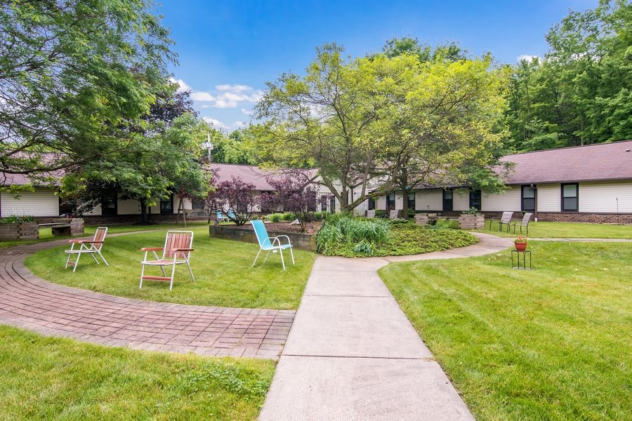 A serene garden setting with chairs and a pathway leading to a building surrounded by lush greenery.