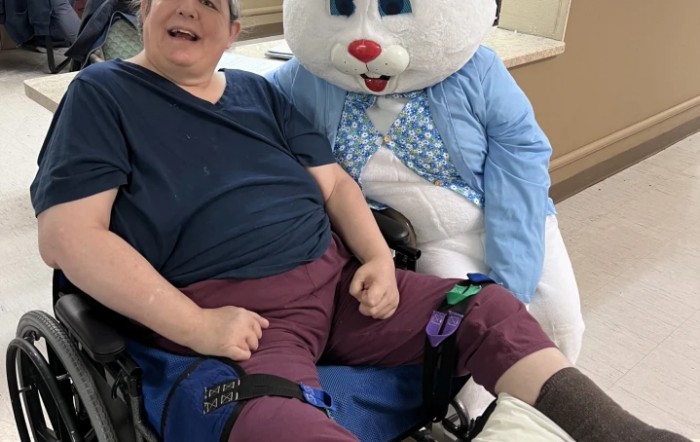 A person in a wheelchair is smiling next to someone dressed as the Easter Bunny indoors.