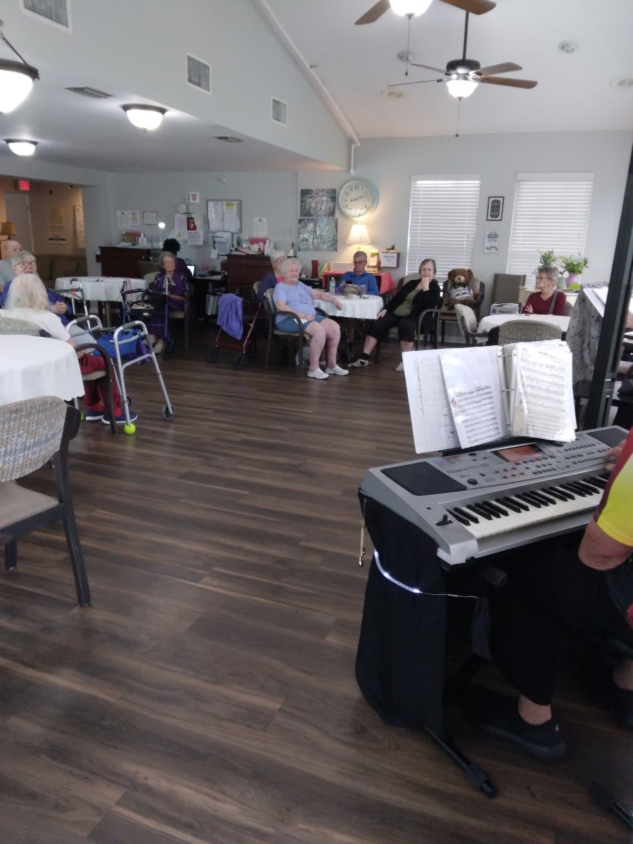 A group of elderly people gathered indoors, enjoying music played on a piano.