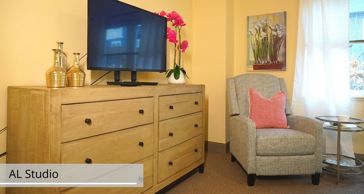 A cozy living room setup featuring a wooden dresser with a television, an armchair, decorative flowers, and wall art.