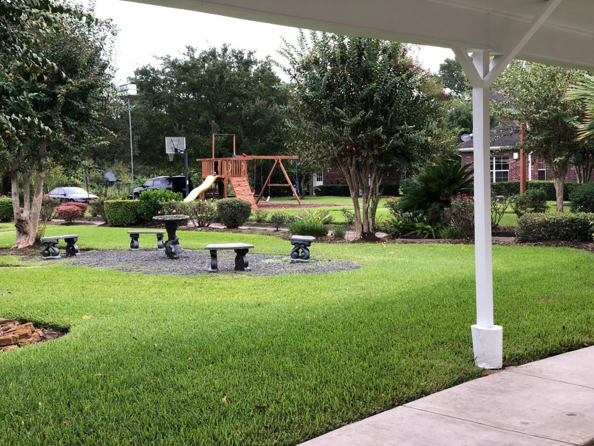 A well-maintained backyard featuring a playground with a swing set and garden benches, surrounded by lush green grass and trees.