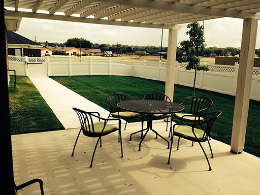 A cozy patio setup with metal table and chairs under a pergola, overlooking a well-maintained lawn and garden.
