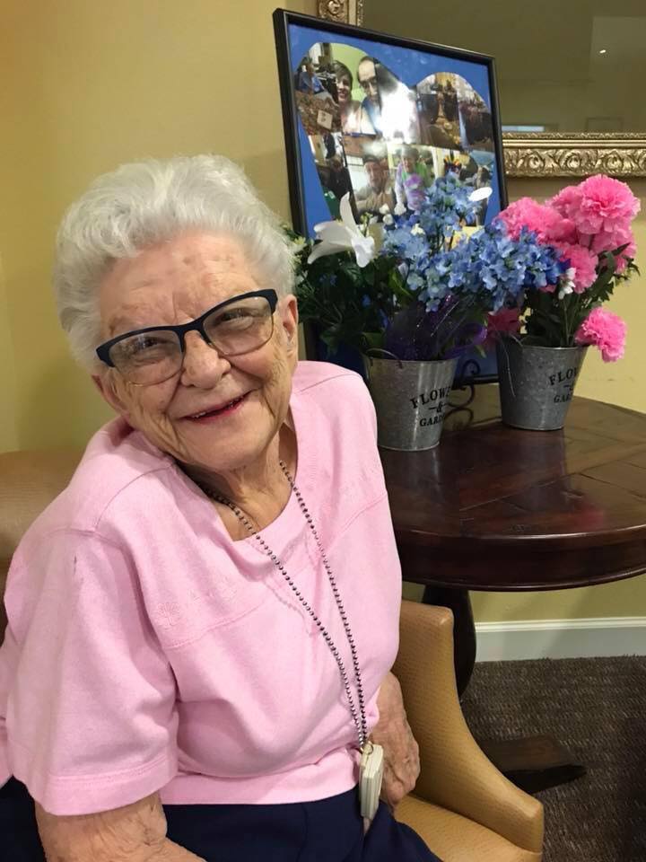An elderly woman with glasses smiles while sitting indoors, with colorful flowers in the background.