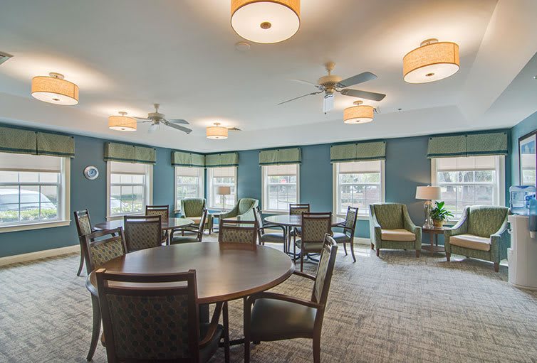A spacious dining room with multiple round tables and chairs, featuring large windows and warm ceiling lighting.