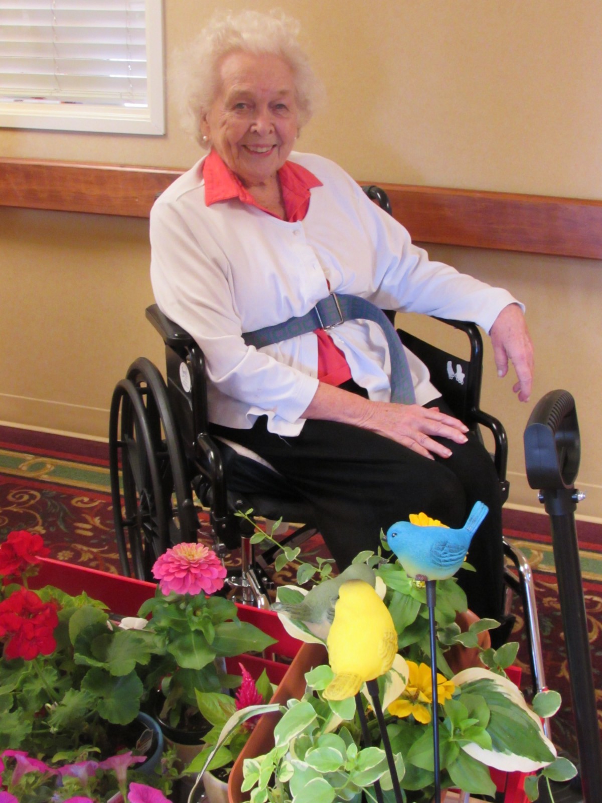 An elderly woman in a wheelchair smiles beside colorful flowers indoors.