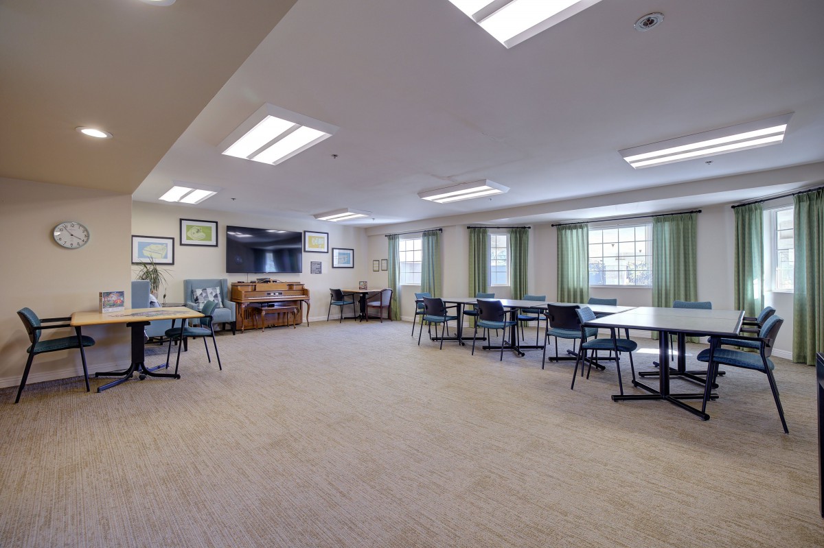 A spacious room with tables and chairs arranged, featuring large windows and a carpeted floor.