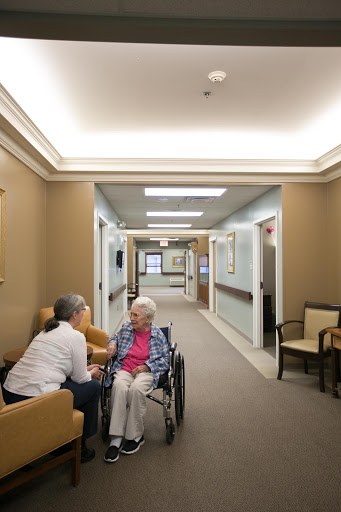 A caregiver attentively interacts with an elderly resident in a well-lit nursing home corridor.