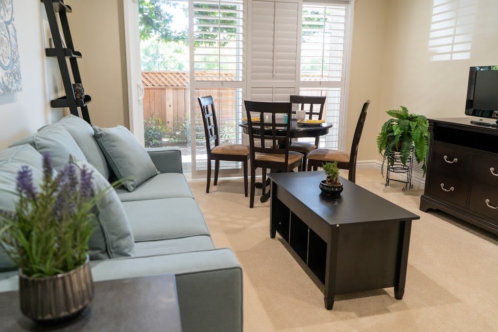 A cozy living room with a sofa, coffee table, and dining area featuring natural light from large windows.