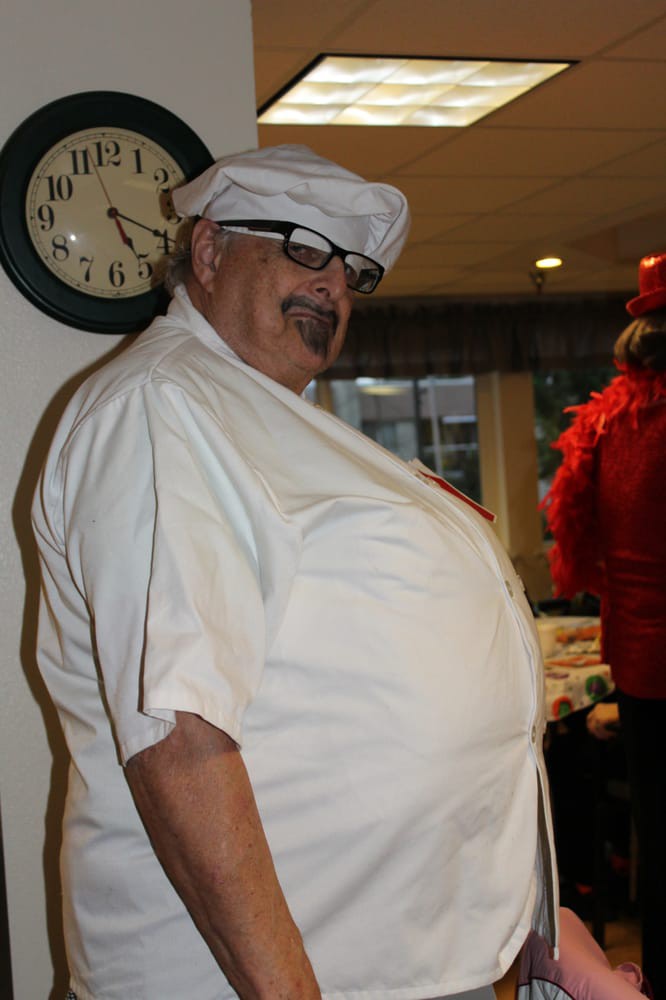 A man wearing a costume with a chef hat and glasses stands indoors, near a clock on the wall.