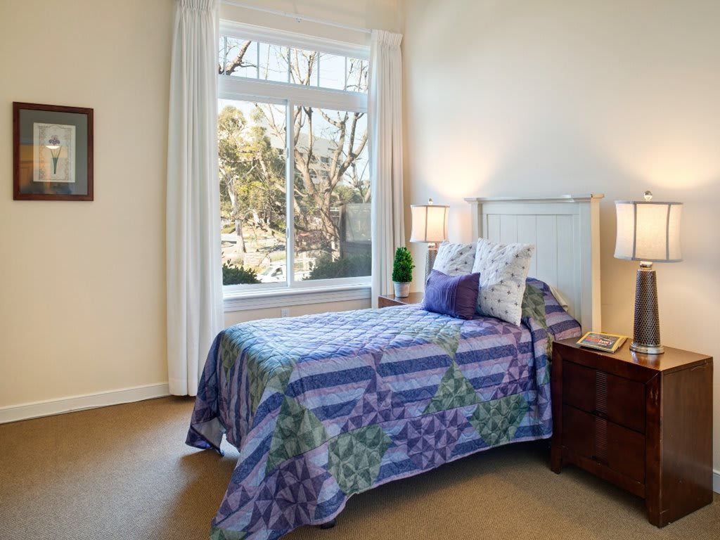 A cozy bedroom with a single bed adorned with colorful bedding, positioned near a large window that lets in natural light. A nightstand with a lamp sits beside the bed.