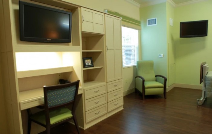 A cozy room interior featuring a desk with a chair, a wall-mounted television, and a wardrobe beside a window.